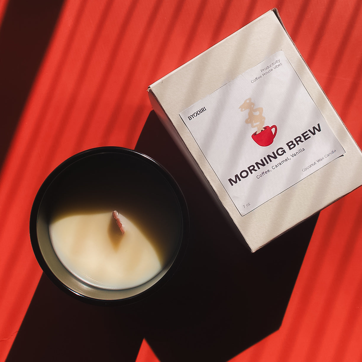 Candle in a black container next to a 'Morning Brew' coffee package on a red surface