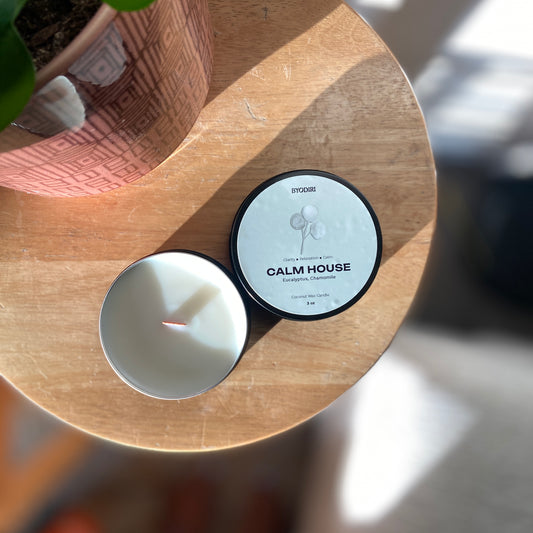 Candle and container labeled 'Calm House' on a wooden surface with a plant in the background.