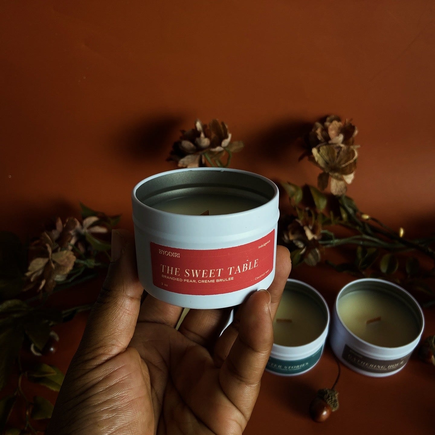 Hand holding a candle labeled 'The Sweet Table' with other candles in the background on a brown surface.