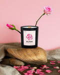 Black candle labeled 'Bloom' with pink roses and petals on a stone surface against a pink background