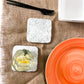 Speckled white concrete coaster set on a burlap table cloth with an orange plate on the right