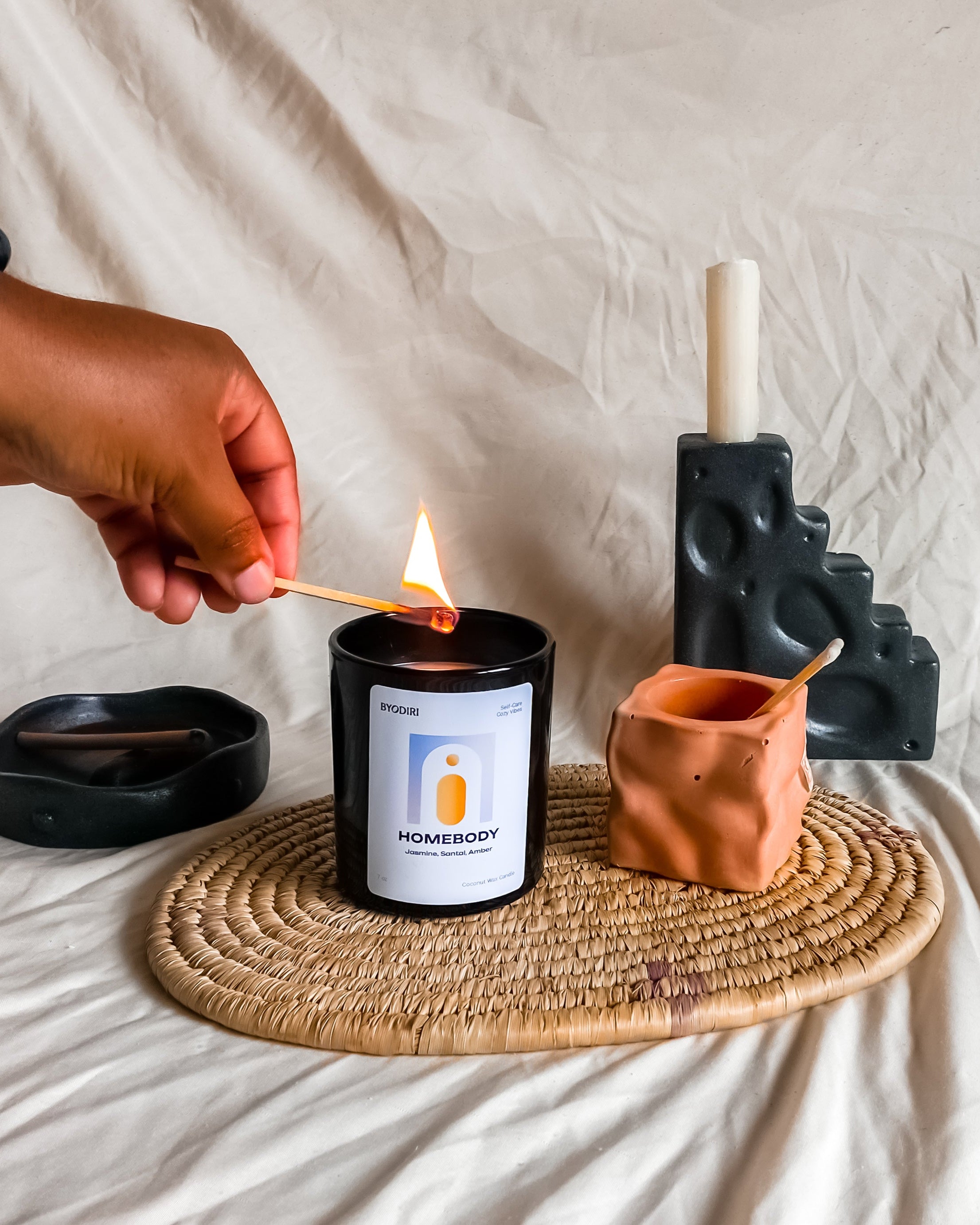 Person lighting a candle with a match on a woven mat, with a neutral background