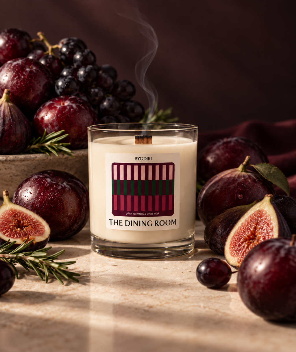A scented candle in a clear glass jar sits on a stone surface surrounded by figs, grapes, rosemary, and a dark draped fabric. The candle wax is creamy off-white, with a printed label reading 'THE DINING ROOM' and a striped graphic in pink, burgundy, and green.
