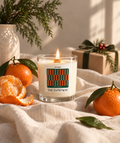 A lit candle in a clear glass jar sits on a cream fabric surface, surrounded by oranges, greenery, and a wrapped gift box. The candle label reads 'BYODIRI' and 'THE ENTRYWAY' with a geometric striped pattern.
