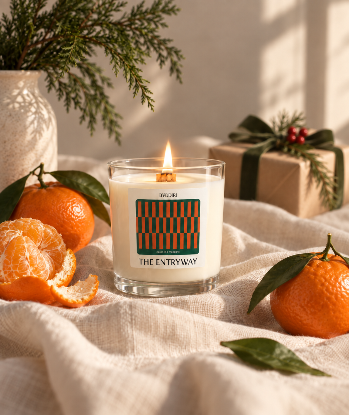 A lit candle in a clear glass jar sits on a cream fabric surface, surrounded by oranges, greenery, and a wrapped gift box. The candle label reads 'BYODIRI' and 'THE ENTRYWAY' with a geometric striped pattern.