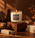 A lit scented candle in a clear glass jar sits on a wooden pedestal in a warm, rustic setting. The candle label reads 'THE SITTING ROOM' with a striped awning graphic.