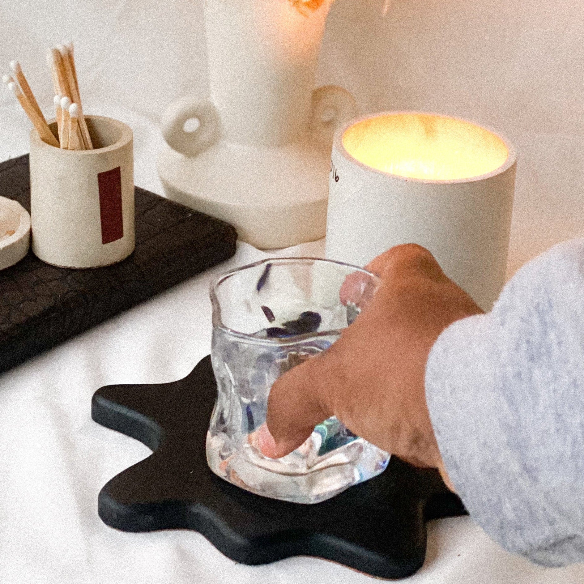 A person's hand lifting a glass off a black concrete coaster on a table, with a candle and other items in the background.