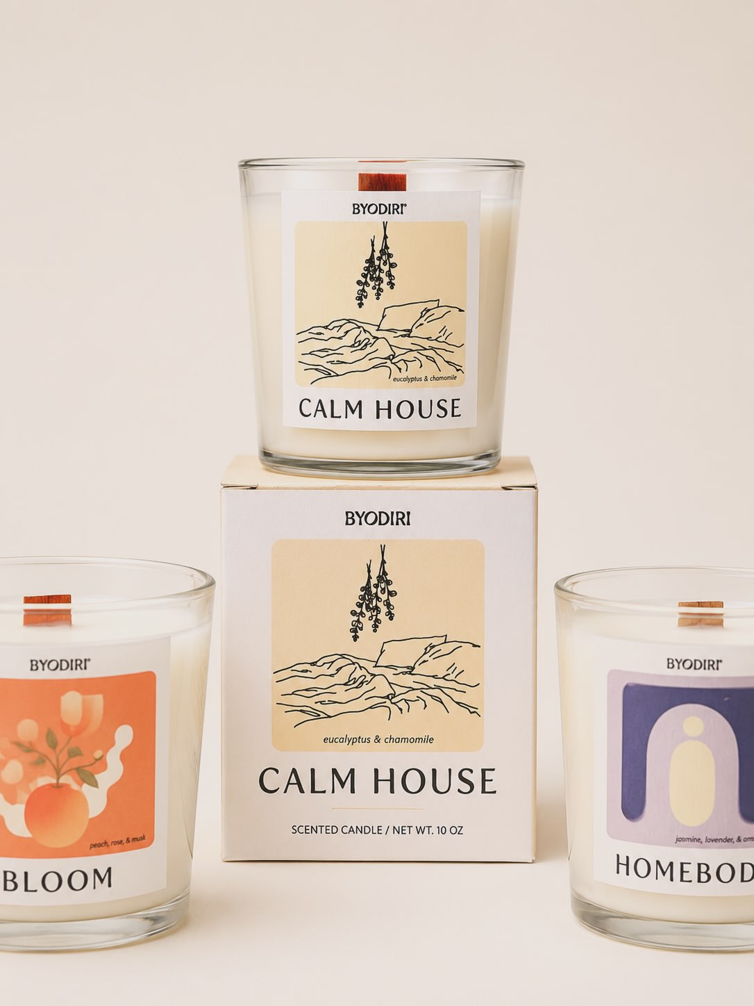 Three BYODIRI scented candles in clear glass jars with cream-colored wax, arranged around a central branded box. The top jar is labeled Calm House, the left jar shows Bloom with an orange abstract design, and the right jar shows Homebody with a lavender arch design.